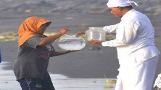 Seorang ibu mengenakan jilbab cokelat rela berbasah-basahan membantu seorang pemangku (pemimpin persembahyangan umat Hindu Bali) mengambil air suci dari pantai. 