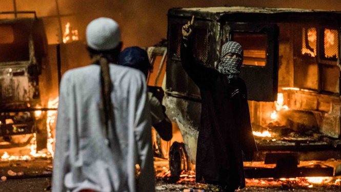 Seorang pengunjuk rasa bercadar mengangkat jari telunjuknya dengan latar belakang mobil yang dibakar dalam kericuhan demo 4 November 2016.