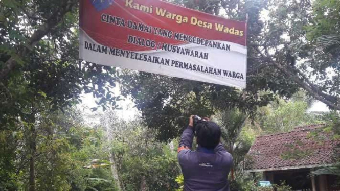 Seorang jurnalis mengabadikan spanduk berisi pesan-pesan penyelesaian konflik secara damai yang terbentang di jalan Desa Wadas, Kecamatan Bener, Kabupaten Purworejo, Jawa Tengah, Selasa, 15 Februari 2022.
